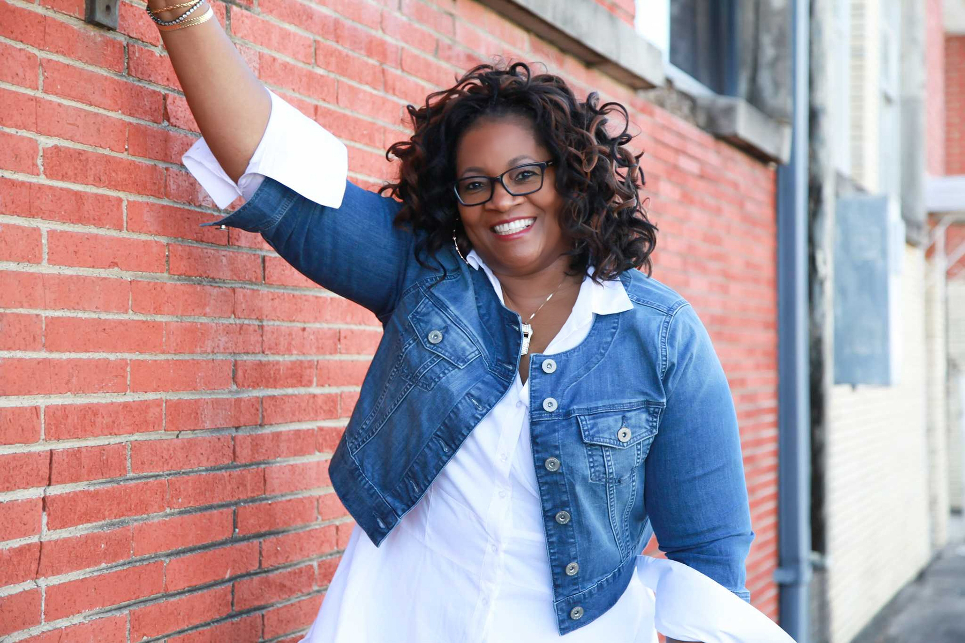 A woman leaning against a brick wall with her arm up.